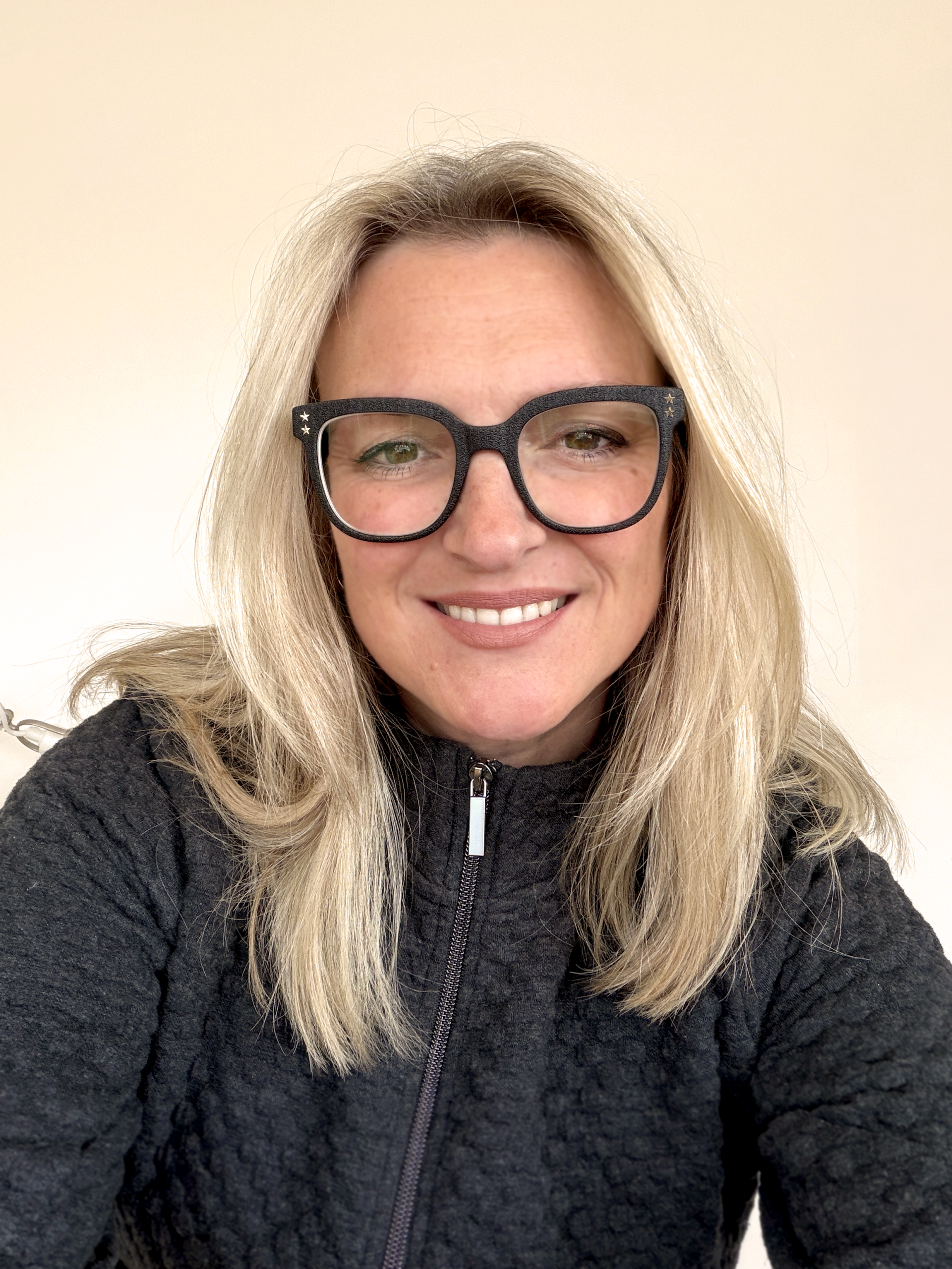 A woman with long blonde hair, wearing black glasses and a dark textured zip-up jacket, smiles at the camera against a plain, light-coloured background.